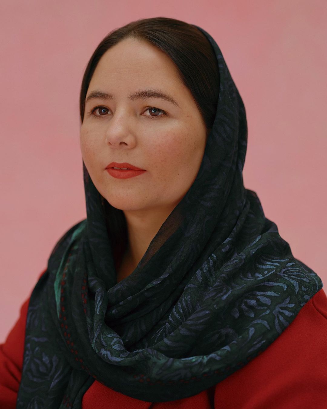 Close-up portrait of a woman with dark hair wearing a patterned headscarf and a red top against a pink background.