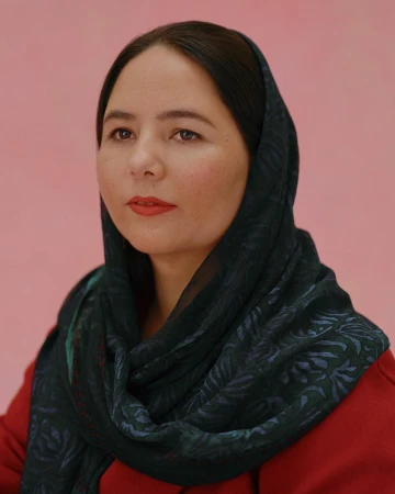 Close-up portrait of a woman with dark hair wearing a patterned headscarf and a red top against a pink background.