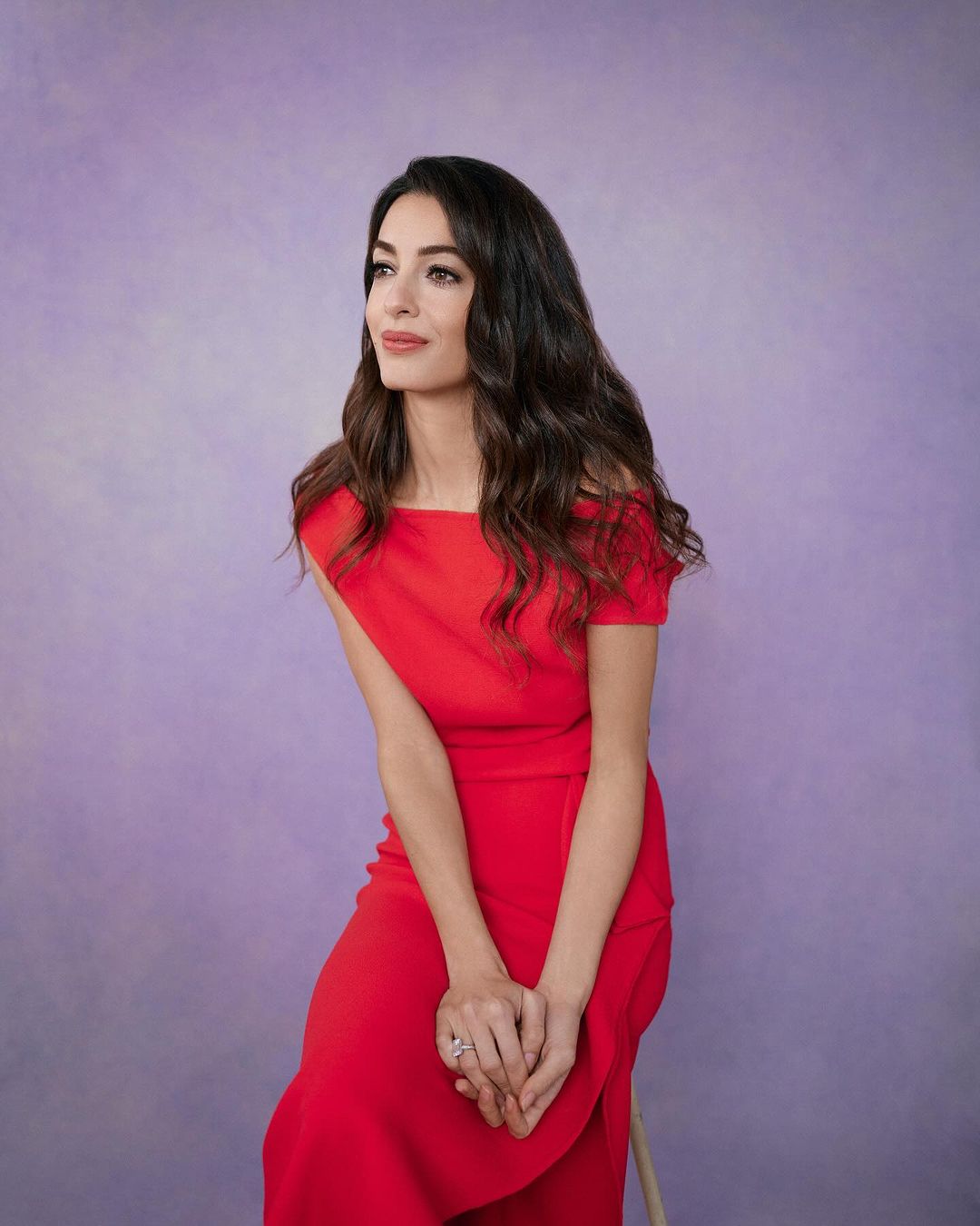 Adult woman in red dress with shoulderless detail sitting in a photo studio with a purple background looking towards the left.