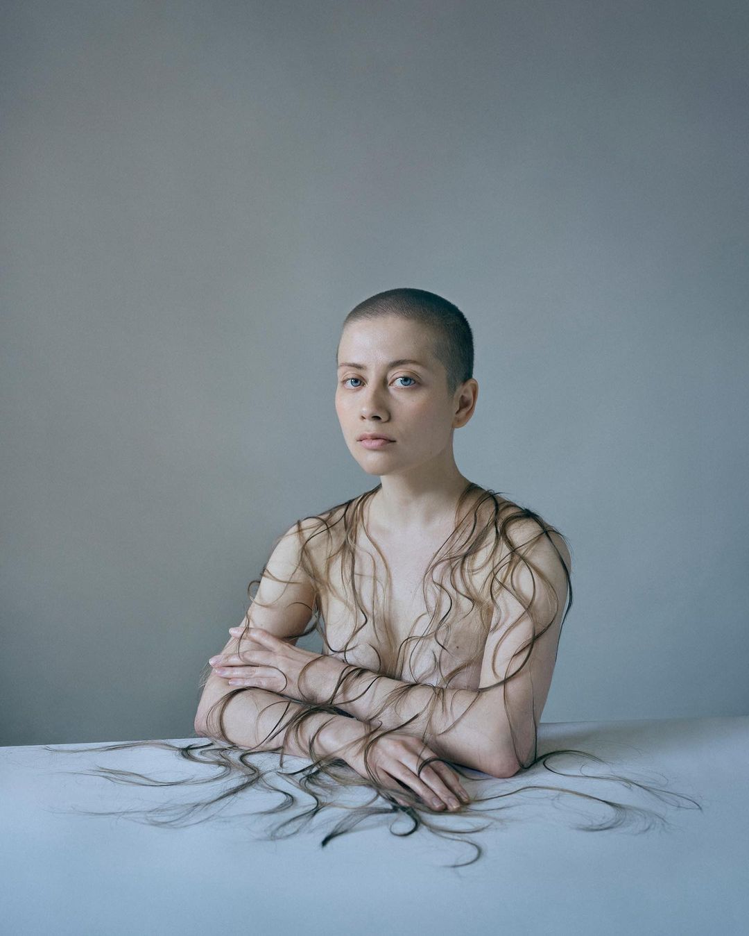 Woman with a shaved head sits at a table with crossed arms, surrounded by strands of dark hair creating a visually striking artistic portrait.