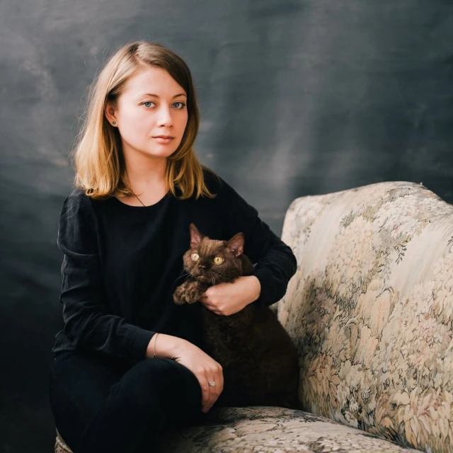 A woman with blonde hair sits on a patterned couch holding a brown cat in her lap. The background is a textured grey and black.