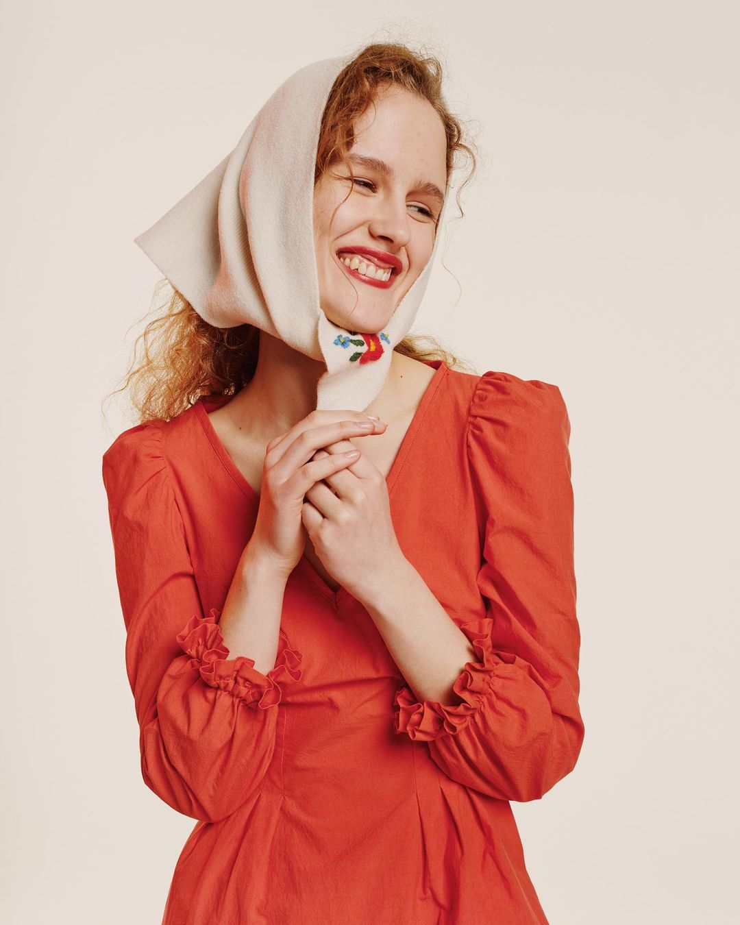 A woman with curly hair smiles, wearing a red dress and a white headscarf with floral embroidery, against a light background.