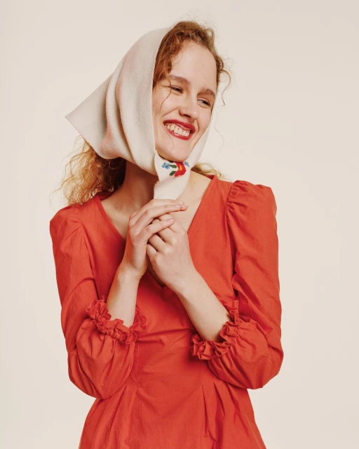 A woman with curly hair smiles, wearing a red dress and a white headscarf with floral embroidery, against a light background.