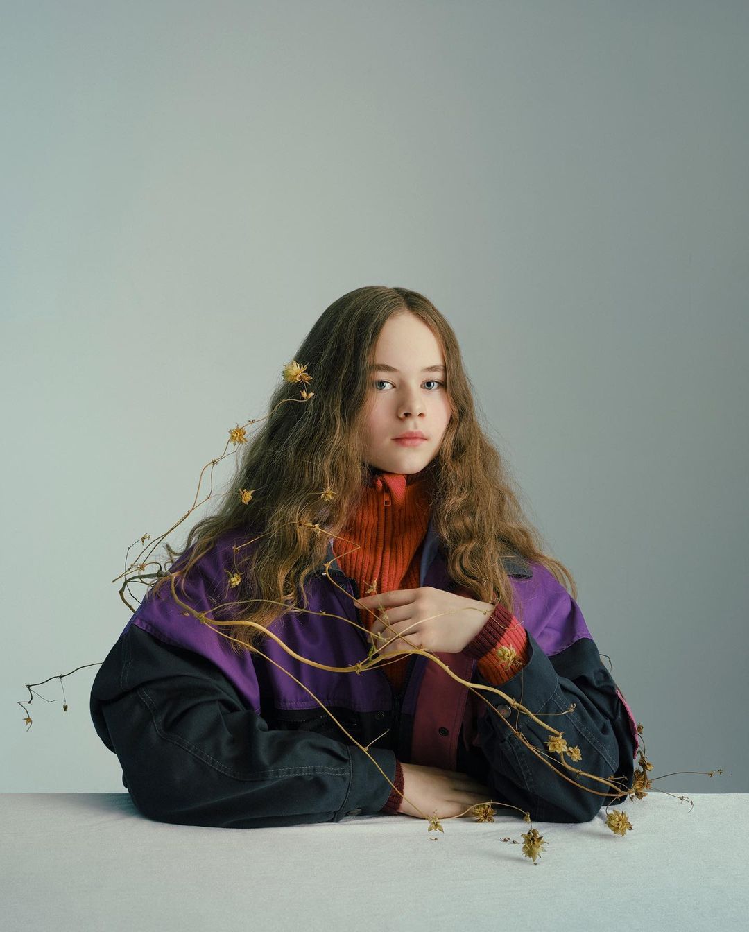 Portrait of a young person with long wavy hair, wearing a color-blocked jacket, indoors in front of a light grey background.
