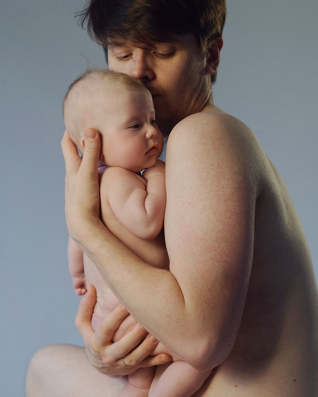 Man holding a baby gently against his chest against a pale grey studio background by photographer Kristina Varaksina
