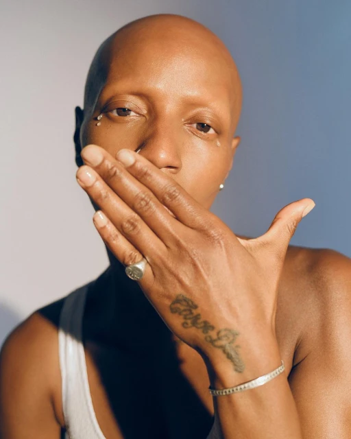 Close-up portrait of a bald Black woman with hand partially covering face, wearing a white tank top and jewelry, against a gradient background.