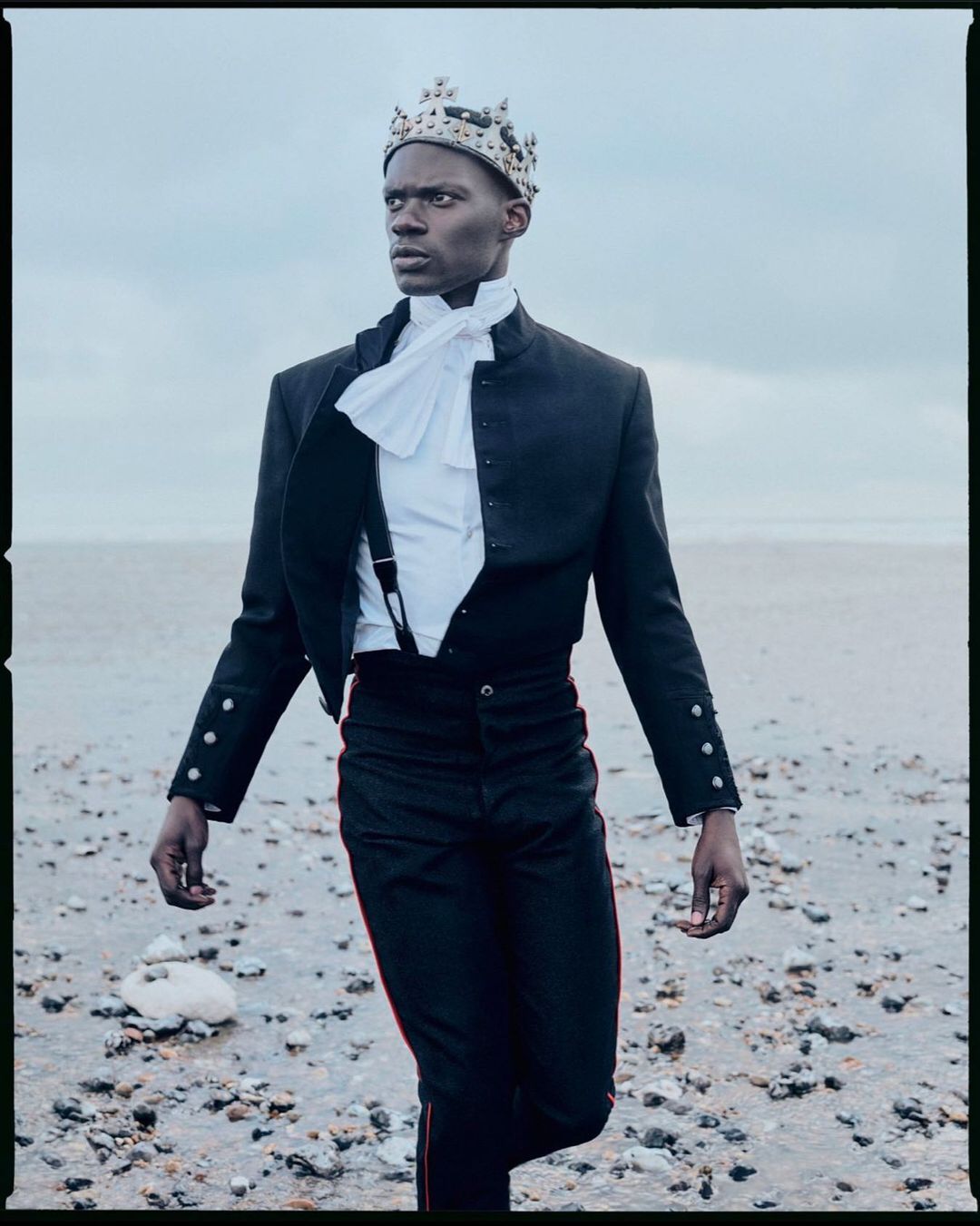 Stylish young Black man wearing a crown, ruffled shirt, suspenders, and fitted jacket on a stony beach under a cloudy sky.