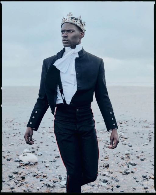 Stylish young Black man wearing a crown, ruffled shirt, suspenders, and fitted jacket on a stony beach under a cloudy sky.