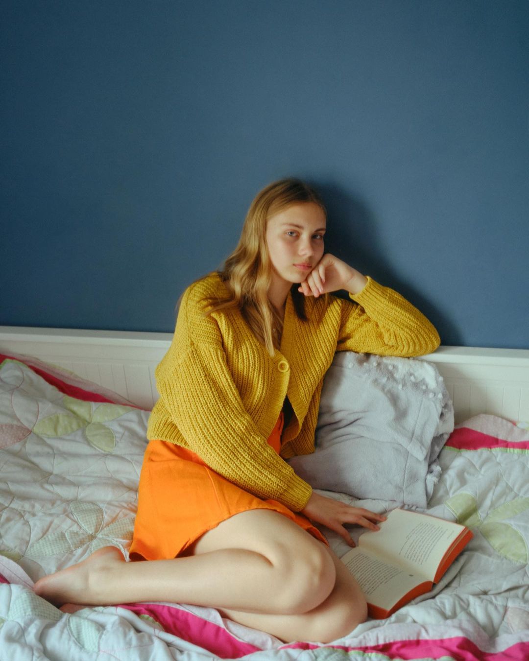 Young woman with blonde hair sits barefoot on a bed with a book, wearing an orange skirt and yellow cardigan against a blue wall.
