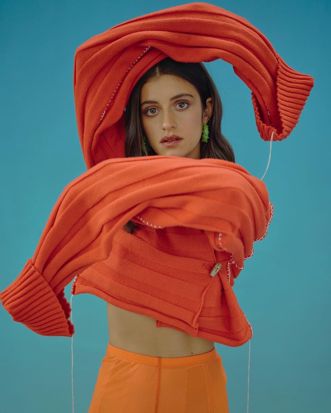 Portrait of a young woman in orange clothing with a red ribbed sweater draped around her against a turquoise background in a photo studio.