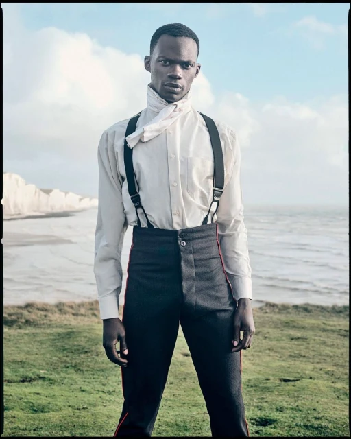Stylish portrait of a Black male model outdoors, wearing a white shirt with black suspenders and high-waisted black pants on a grassy hill.