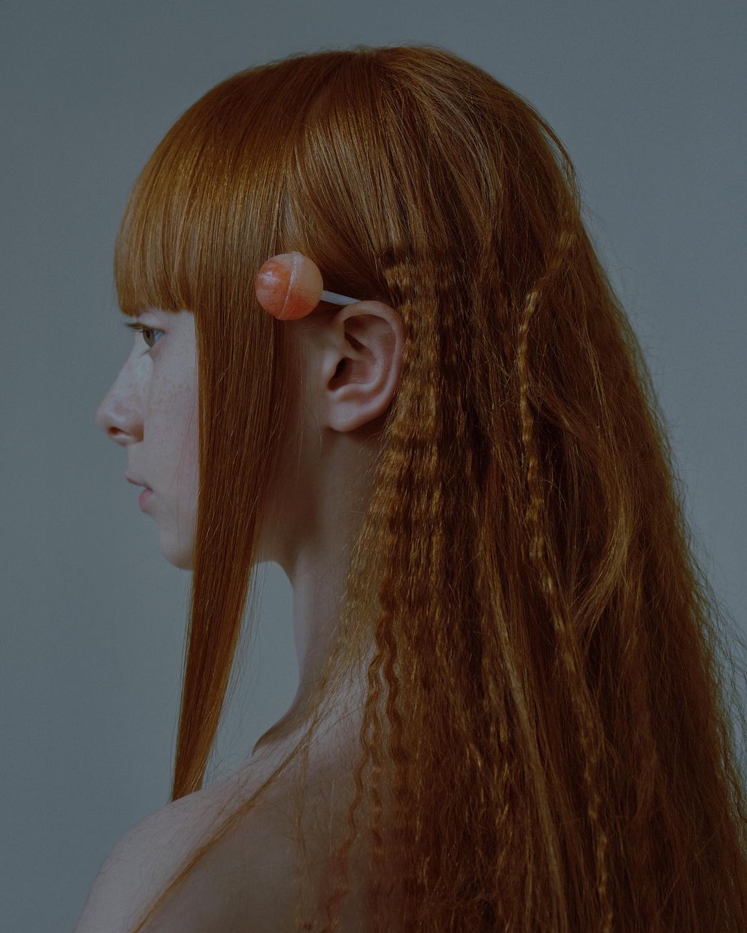 Profile portrait of a young woman with auburn hair, styled with crimped sections and a lollipop accent, set against a solid light grey background.