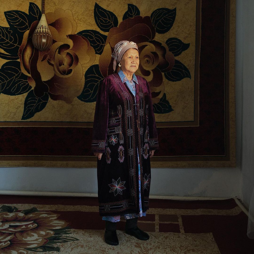 Elderly woman in embroidered coat stands in room with ornate wall and floor rugs in earth tones, wearing headscarf and dark shoes.