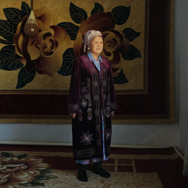 Elderly woman in embroidered coat stands in room with ornate wall and floor rugs in earth tones, wearing headscarf and dark shoes.