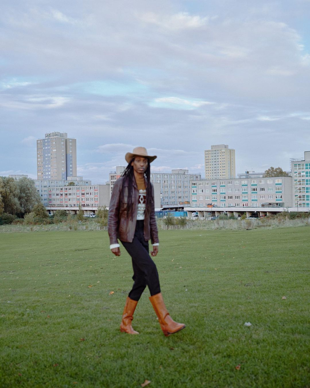 A person in a brown jacket, cowboy hat, and boots walks across a green field with urban buildings in the background under a cloudy sky.