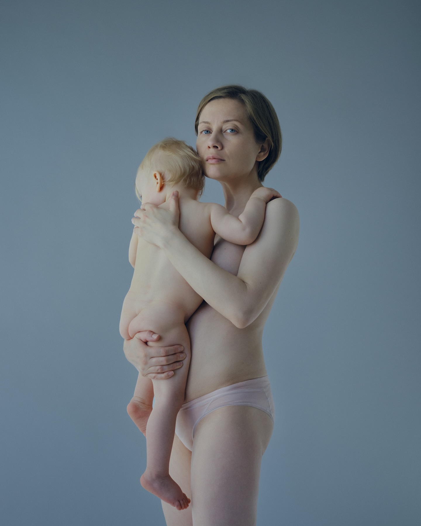A mother with short hair gently holds her baby in a studio setting, both partially undressed against a light blue background.