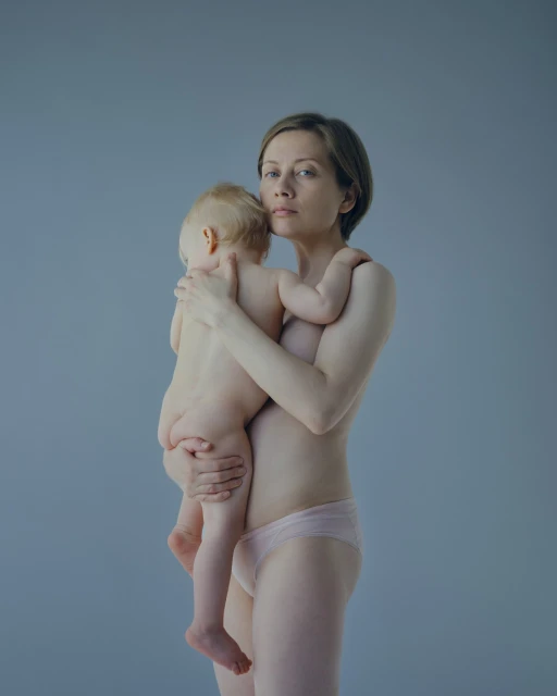 A mother with short hair gently holds her baby in a studio setting, both partially undressed against a light blue background.