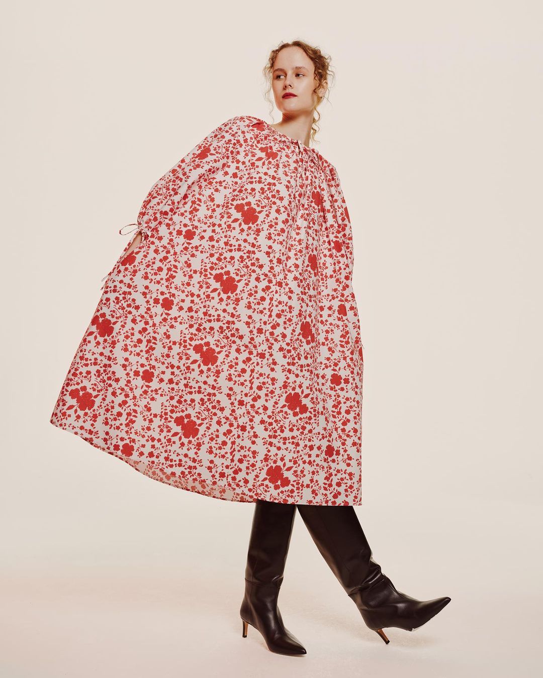 Model in voluminous red and white floral dress wearing black leather boots against a white studio background