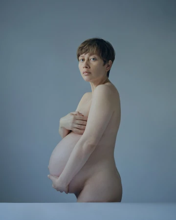A pregnant, nude woman stands in a portrait, facing the camera with a solemn expression, her hands gently supporting her baby bump.