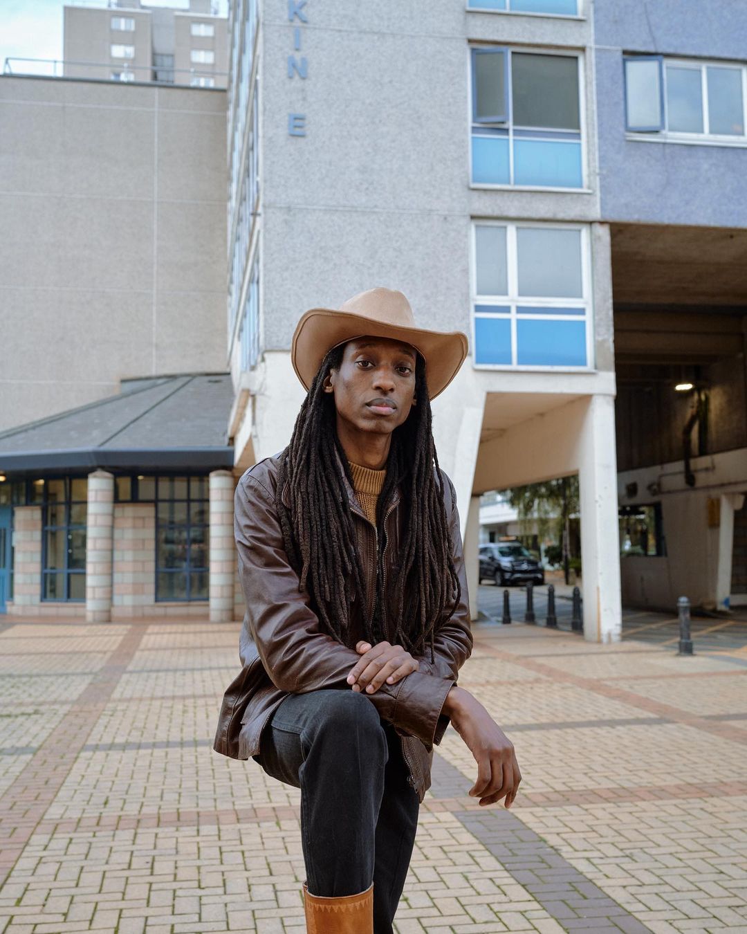 Portrait of a Black adult male with dreadlocks, wearing a cowboy hat and leather jacket, posing in an urban environment with buildings.