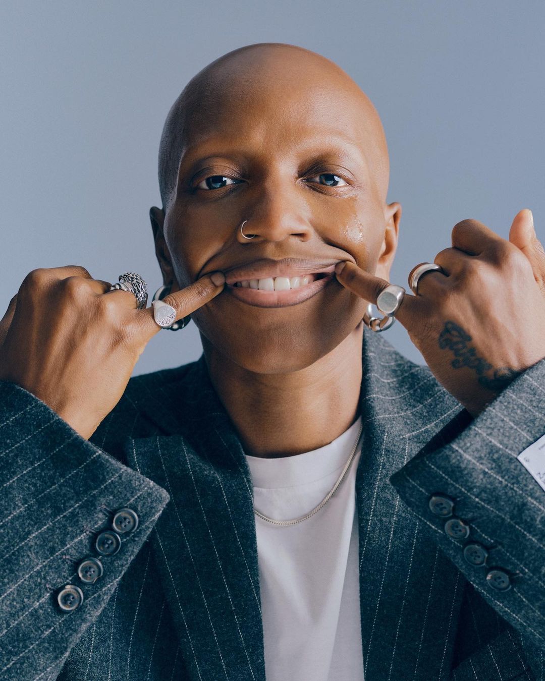 Close-up of a bald adult with a nose ring and a striped blazer pulling their lips into a smile with their fingers in a studio setting.
