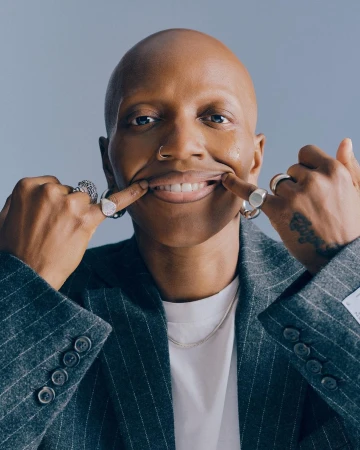 Close-up of a bald adult with a nose ring and a striped blazer pulling their lips into a smile with their fingers in a studio setting.