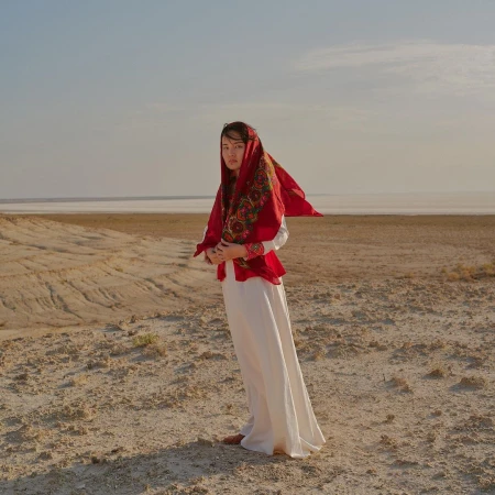 Woman in white dress and red headscarf standing in a barren desert landscape under an open sky