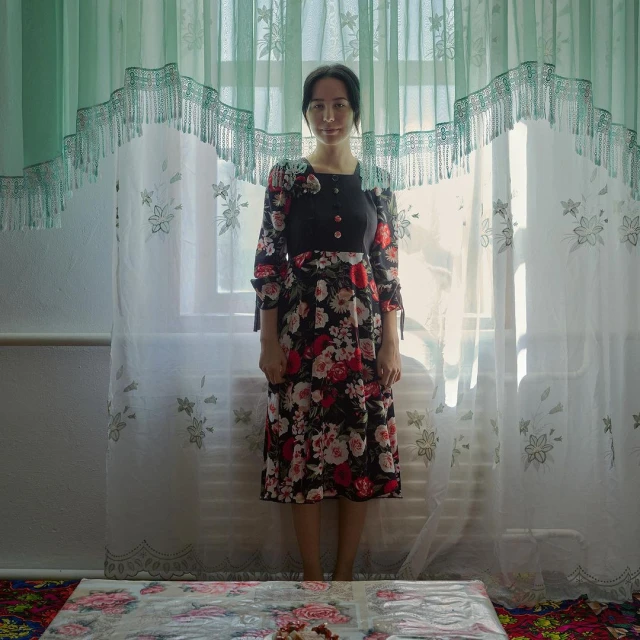 A woman stands in front of a window with floral curtains, wearing a floral dress with long sleeves and a dark top.