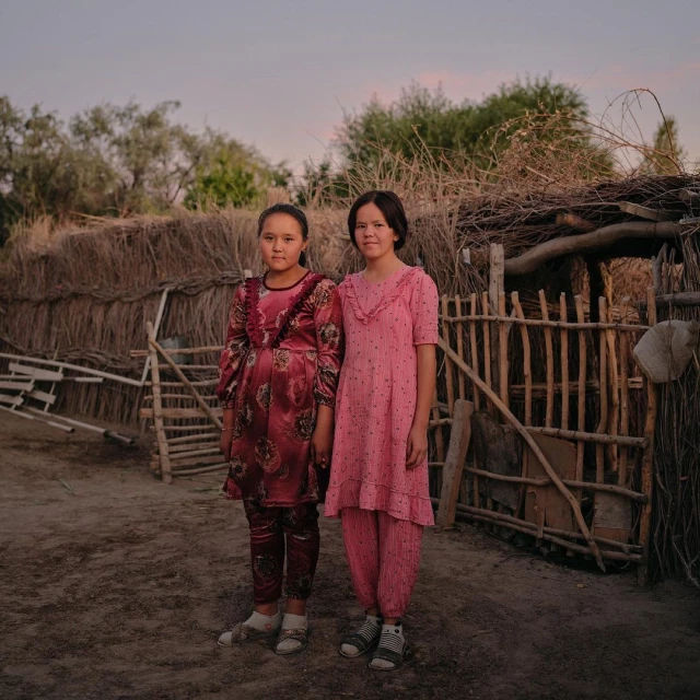Two young girls pose together in an outdoor setting, wearing matching casual dresses and pants, standing in front of a wattle fence.
