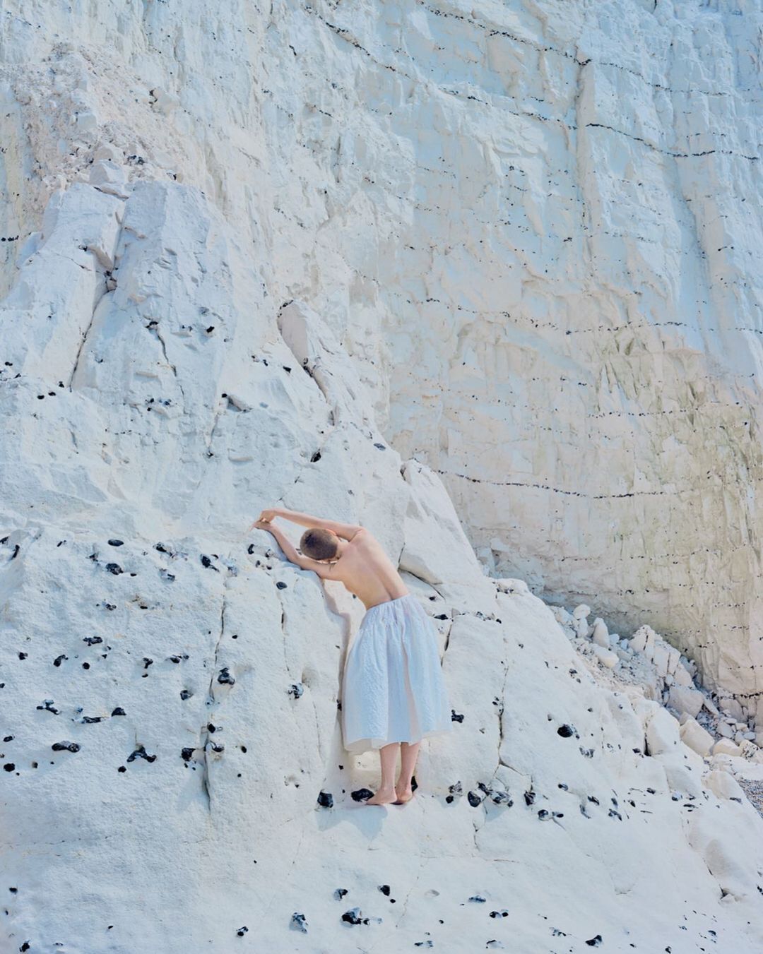 A woman in a white dress stands barefoot against a white cliff face with dark markings in a scenic outdoor setting. The light is bright.