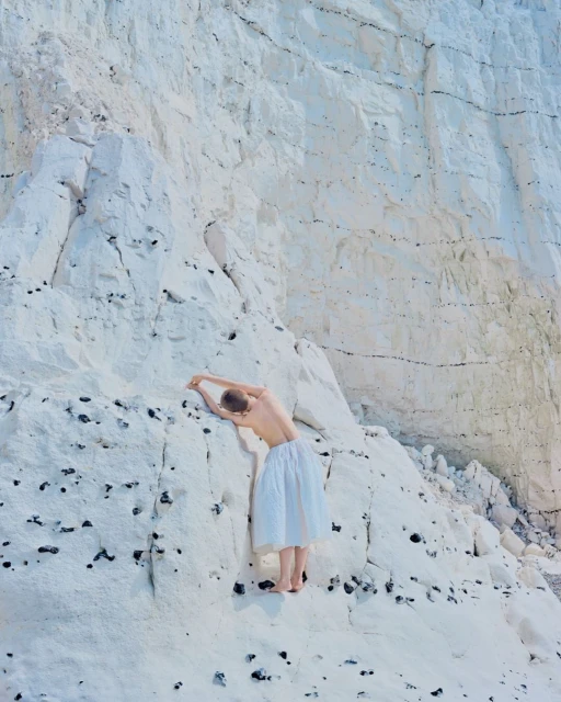 A woman in a white dress stands barefoot against a white cliff face with dark markings in a scenic outdoor setting. The light is bright.