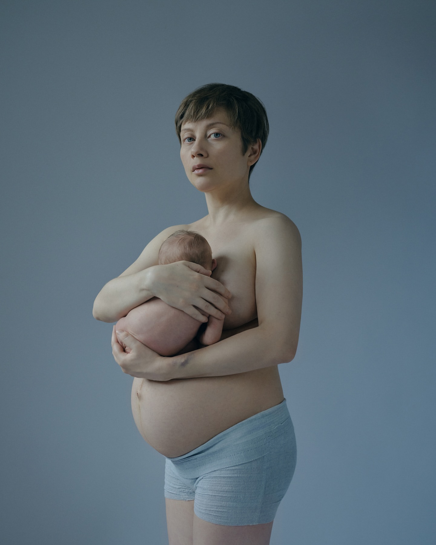 Portrait of a fair-skinned pregnant woman holding a newborn baby against a blue-gray background in a studio setting with soft lighting.