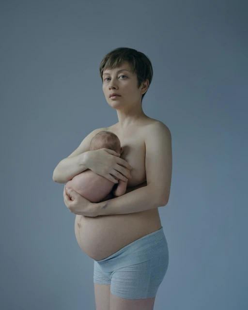 Portrait of a fair-skinned pregnant woman holding a newborn baby against a blue-gray background in a studio setting with soft lighting.