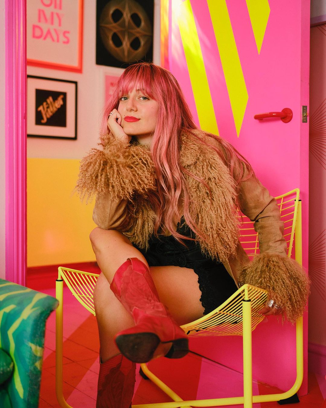 A woman with pink hair poses in a yellow chair, wearing a fur jacket and red boots, in a room with pink and yellow decor.