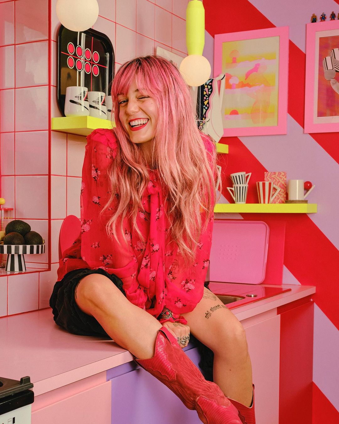 Cheerful woman with pink hair wearing a red floral blouse and boots sits in a vibrant, retro-styled pink and red kitchen.