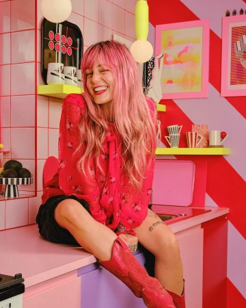 Cheerful woman with pink hair wearing a red floral blouse and boots sits in a vibrant, retro-styled pink and red kitchen.