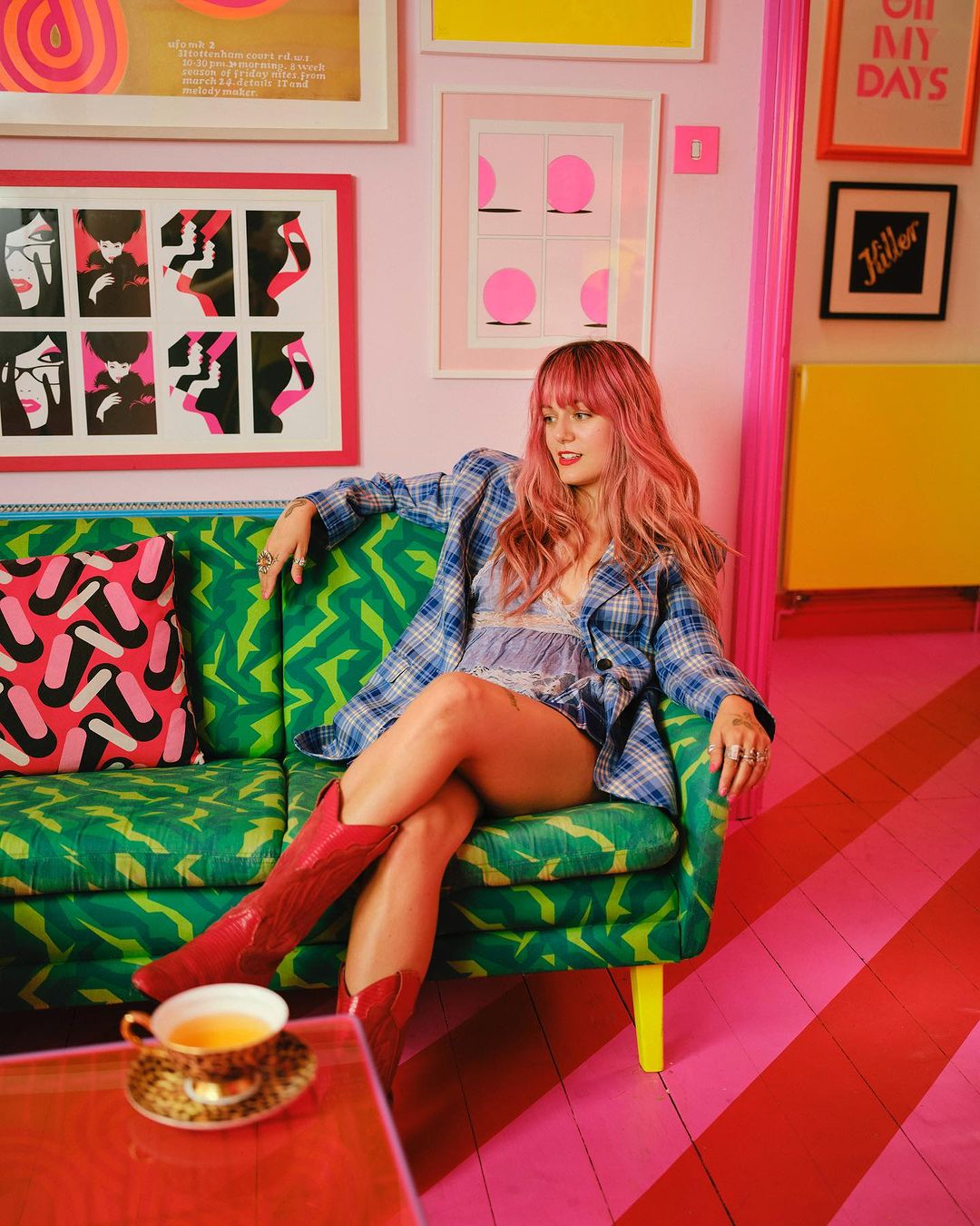 A woman with pink hair sits on a green patterned sofa in a pink room, wearing a blue plaid shirt and red boots, with colorful artwork.