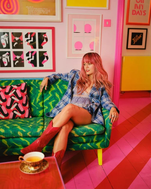 A woman with pink hair sits on a green patterned sofa in a pink room, wearing a blue plaid shirt and red boots, with colorful artwork.