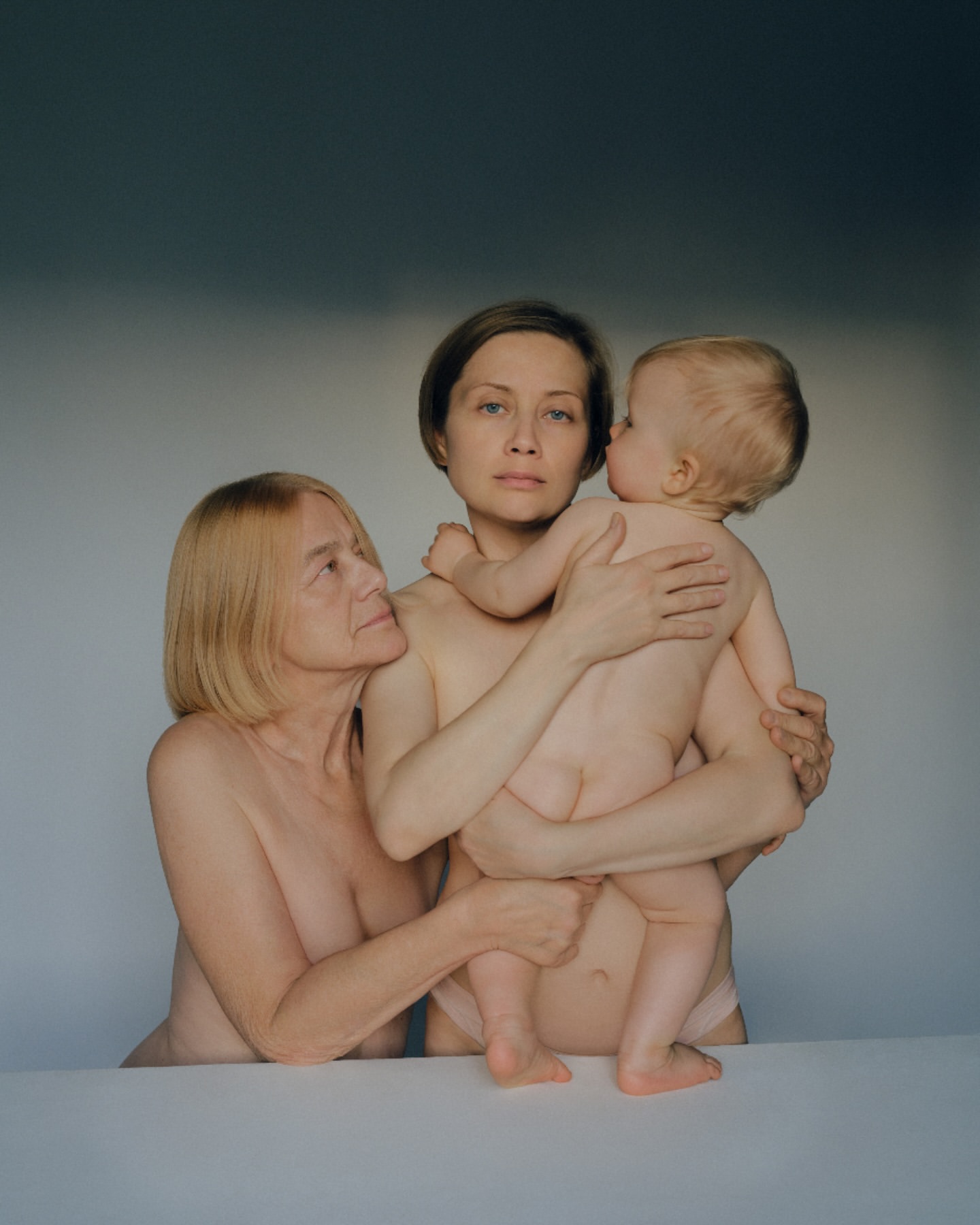 Candid portrait of three generations of women: an elderly woman, a middle-aged woman holding a baby, and a neutral gray background.