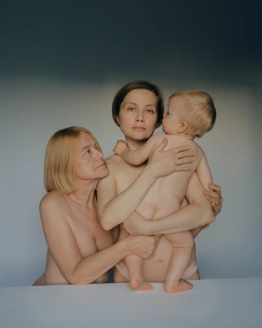 Candid portrait of three generations of women: an elderly woman, a middle-aged woman holding a baby, and a neutral gray background.