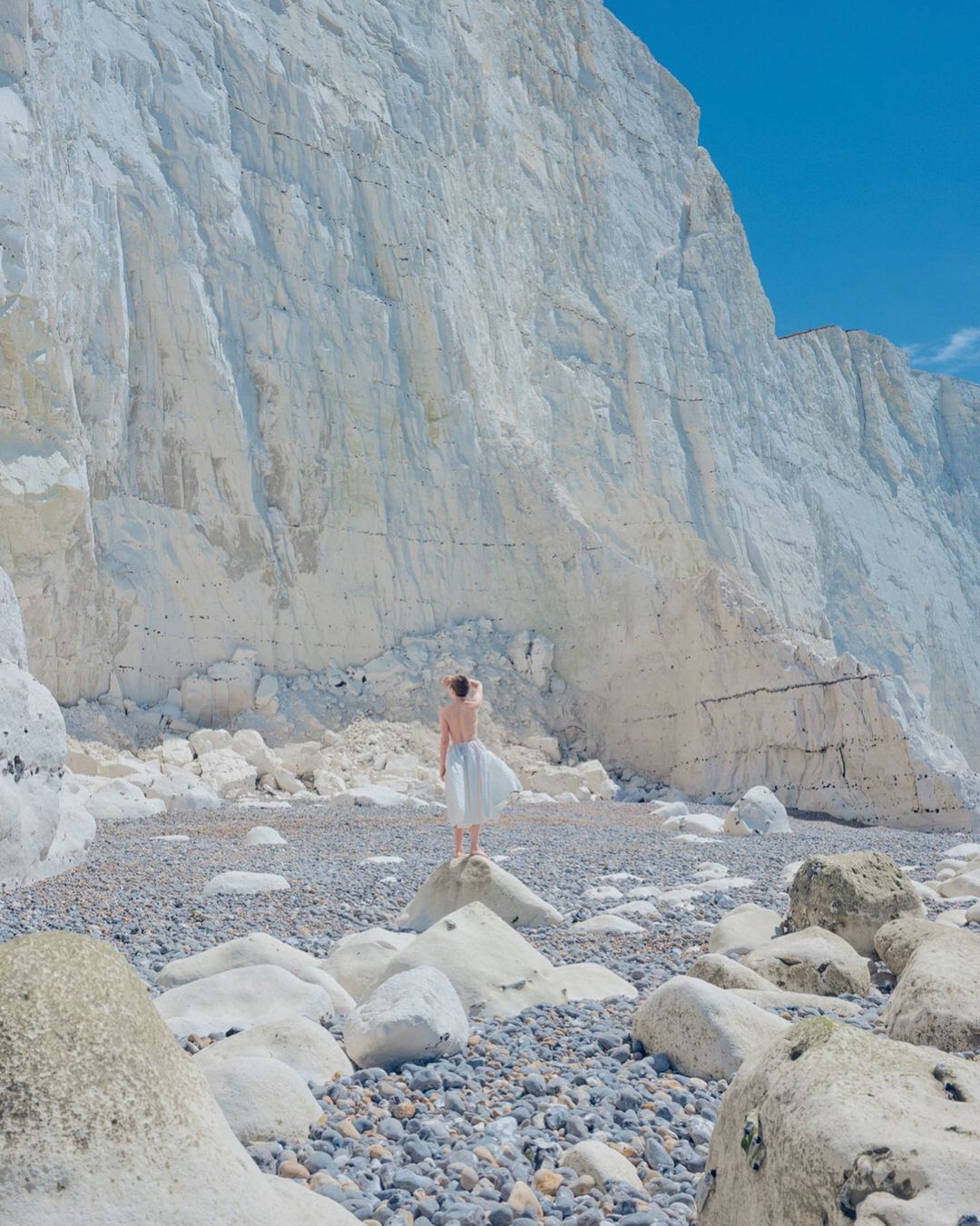 Woman in flowing dress stands on rocks near chalk cliffs under blue sky. Coastal scene showcases nature's beauty and serene landscape.