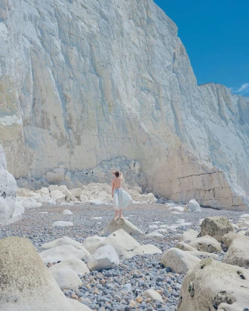 Woman in flowing dress stands on rocks near chalk cliffs under blue sky. Coastal scene showcases nature's beauty and serene landscape.