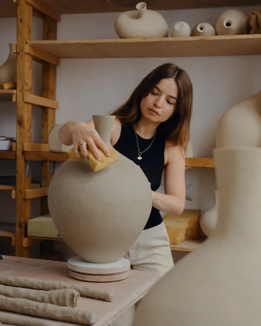 Adult female shaping a large clay vase in a studio; she holds a sponge to smooth the vase surface. Wooden shelves in background.