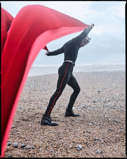 Male model on a beach wearing a crown, black outfit with red trim, and black boots holds a large red fabric against an ocean backdrop.
