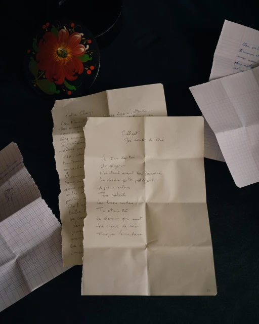 Close-up still life features handwritten letters, a decorative floral container, arranged on a dark surface, suggesting intimacy and reflection.