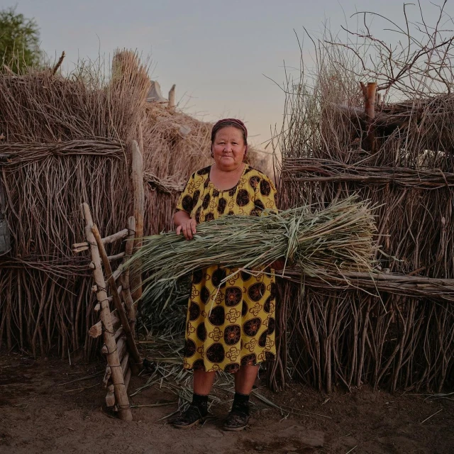 Elderly woman in patterned dress standing outdoors holding bundle of green foliage in front of a rough-hewn stick fence.