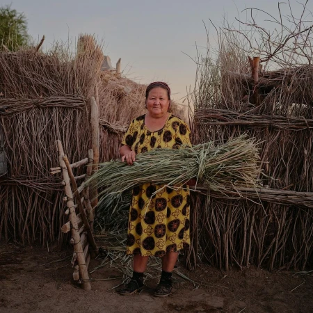 Elderly woman in patterned dress standing outdoors holding bundle of green foliage in front of a rough-hewn stick fence.