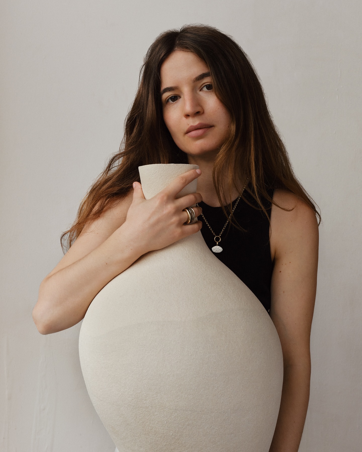Portrait of a woman with long brown hair, wearing a black top and gold necklace and rings, embracing a large, light beige ceramic vase indoors.