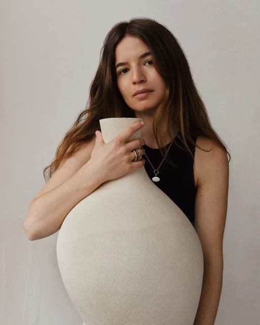 Portrait of a woman with long brown hair, wearing a black top and gold necklace and rings, embracing a large, light beige ceramic vase indoors.