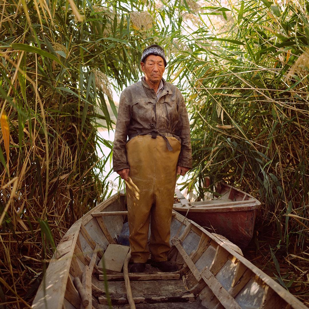 Elderly Asian fisherman in waders stands in a wooden boat surrounded by tall reeds, reflecting a life on the water in a natural setting.